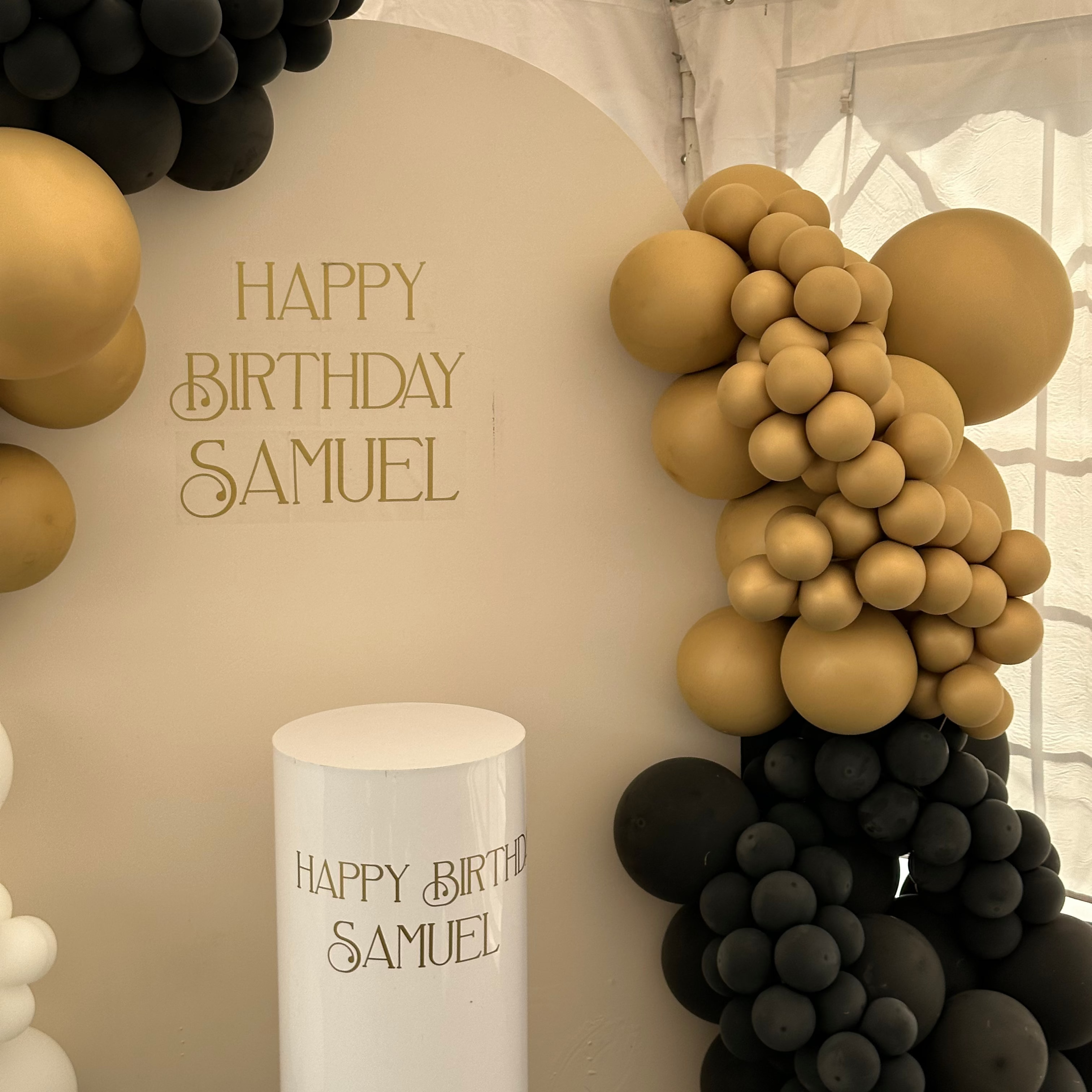 Custom birthday party backdrop setup featuring a white arch with gold 'HAPPY BIRTHDAY SAMUEL' lettering, decorated with black and gold balloon garlands, and a matching white cake stand with personalized text.