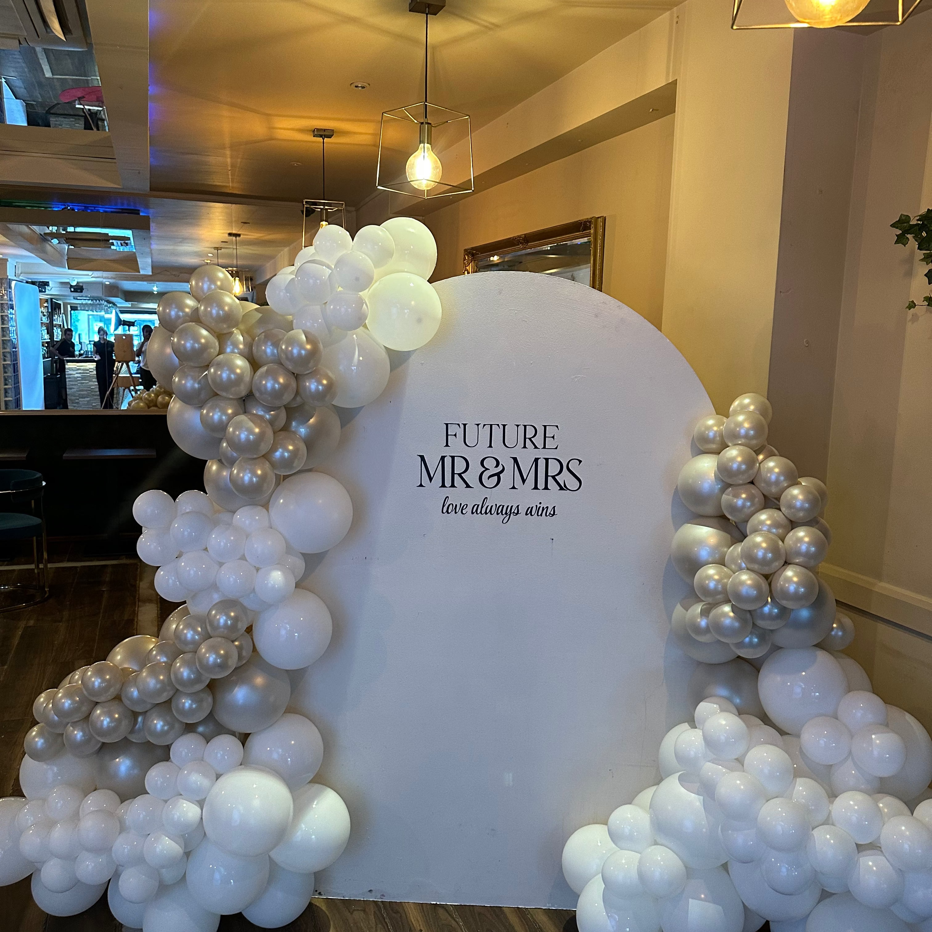 A decorative arch setup for a wedding or event, featuring a large white backdrop with the text 'FUTURE MR & MRS' and 'love always starts here,' surrounded by clusters of white, silver, and pearl balloons in a warmly lit indoor venue.