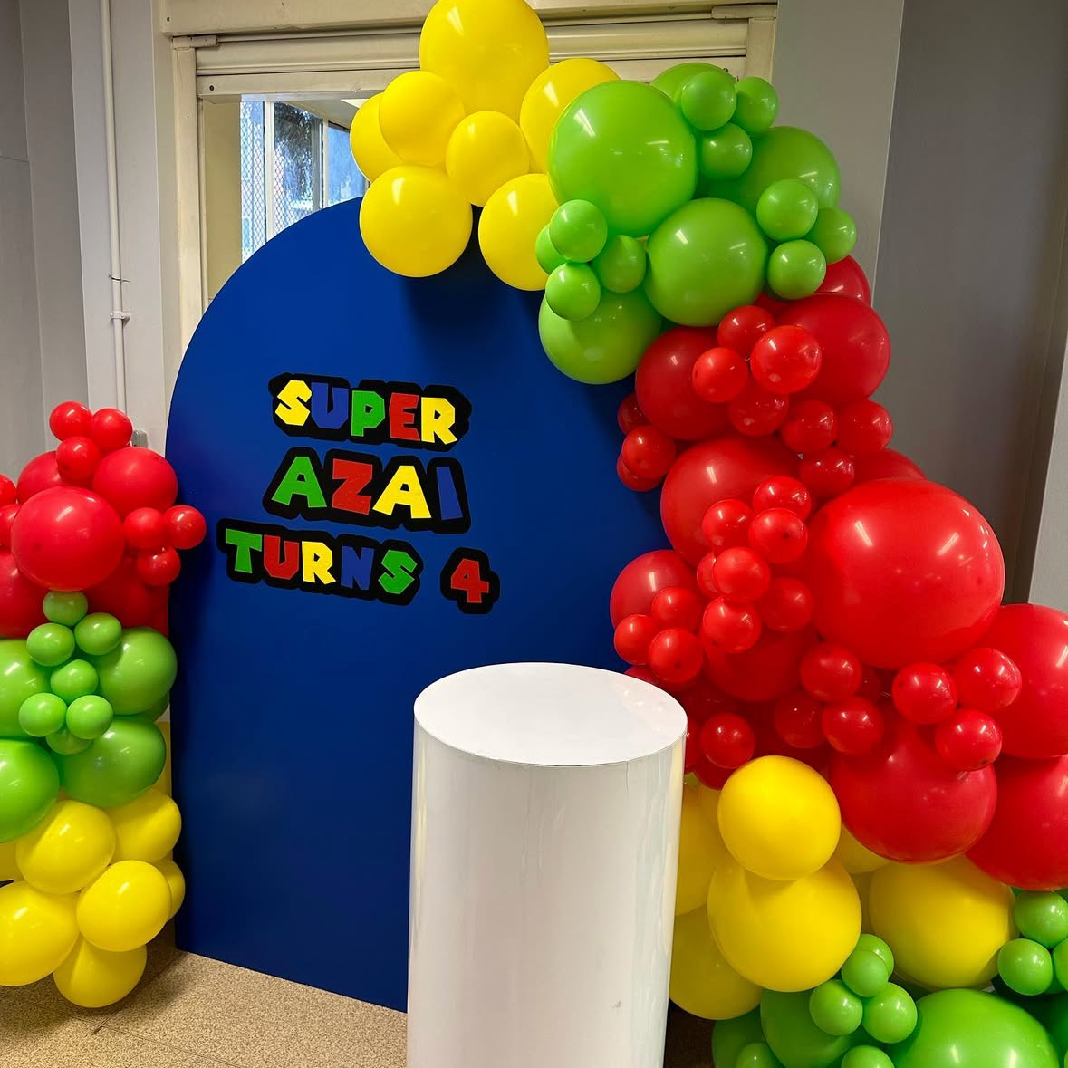 A colorful party backdrop setup with a large blue arch background, decorated with red, yellow, and green balloon clusters. The words 'SUPER AZAI TURNS 4' are displayed in large, colorful letters on the blue backdrop, and a white cylindrical pedestal stands in front. Mario kart edition